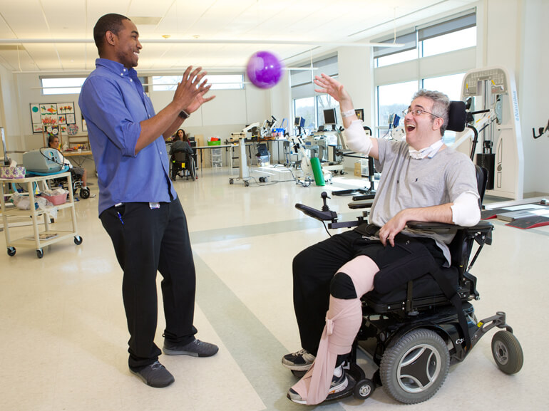 Male patient in wheelchair tossing small purple ball to a therapist.