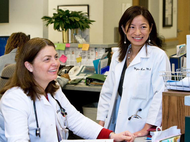 Two nurses in white lab coats at nursing desk in hospital.