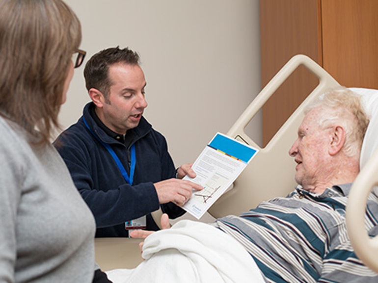 Healthcare professional discussing a care plan with a patient in bed and a caregiver standing nearby.