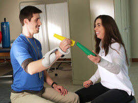 Male patient wearing arm brace putting a yellow tube over a green tube held by therapist.
