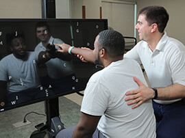 Male therapist works with male patient pointing to letters on a large TV monitor.