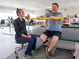 Male patient in therapy gym holding a yellow tube in outstretched arms.