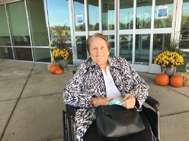 Arlene in wheelchair outside hospital entrance with fall flowers and pumpkins behind her.