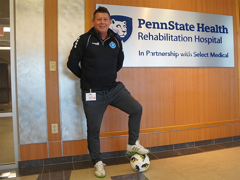 Denis standing in hospital hallway with one foot resting on a soccer ball.