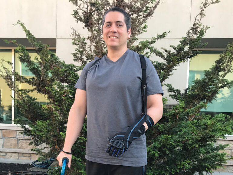 Isaac wearing black robotic arm brace and using a cane while standing outside.