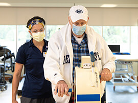 Older male patien wearing Penn State hat and jacket using small motorized arm exerciser.
