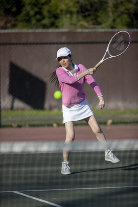 Female tennis player in pink jersey and white skorts making a back swing on the court.