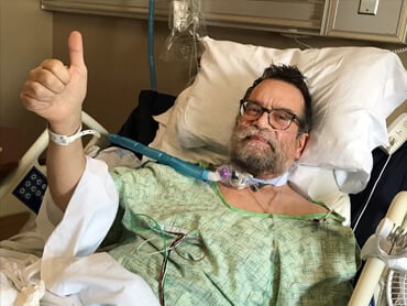 Gregg Wysocki lays in his hospital bed, utilizing a ventilator to breathe, while giving the camera a thumbs up.