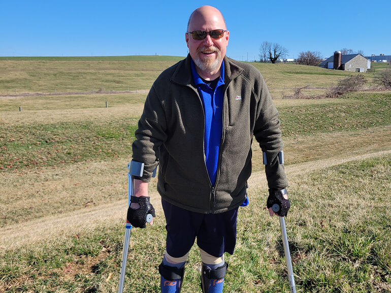 Dane Lauver smiles from behind sunglasses as he stands in a field, holding himself up with crutches.