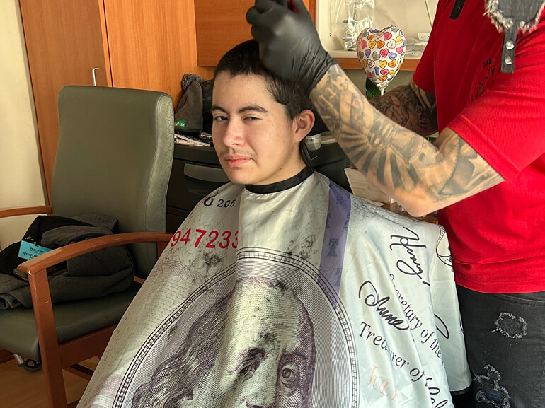 Leo Hernandez gets a haircut under a cape. The barber has tattoos and wears gloves.