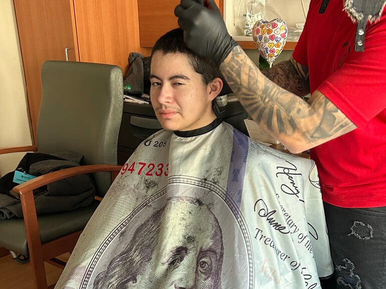 Leo Hernandez gets a haircut under a cape. The barber has tattoos and wears gloves.