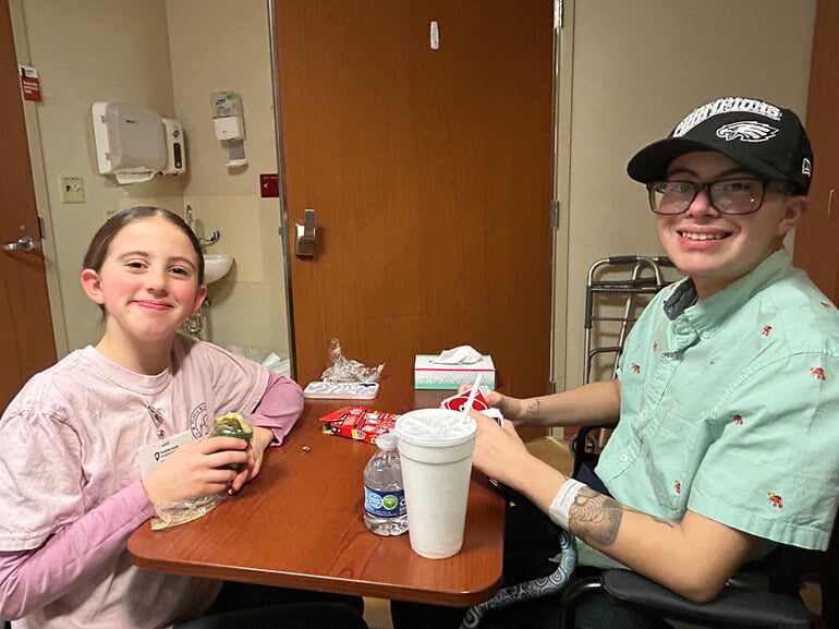 Leo sits at a table in a hospital room with his younger sister. He wears a baseball cap.